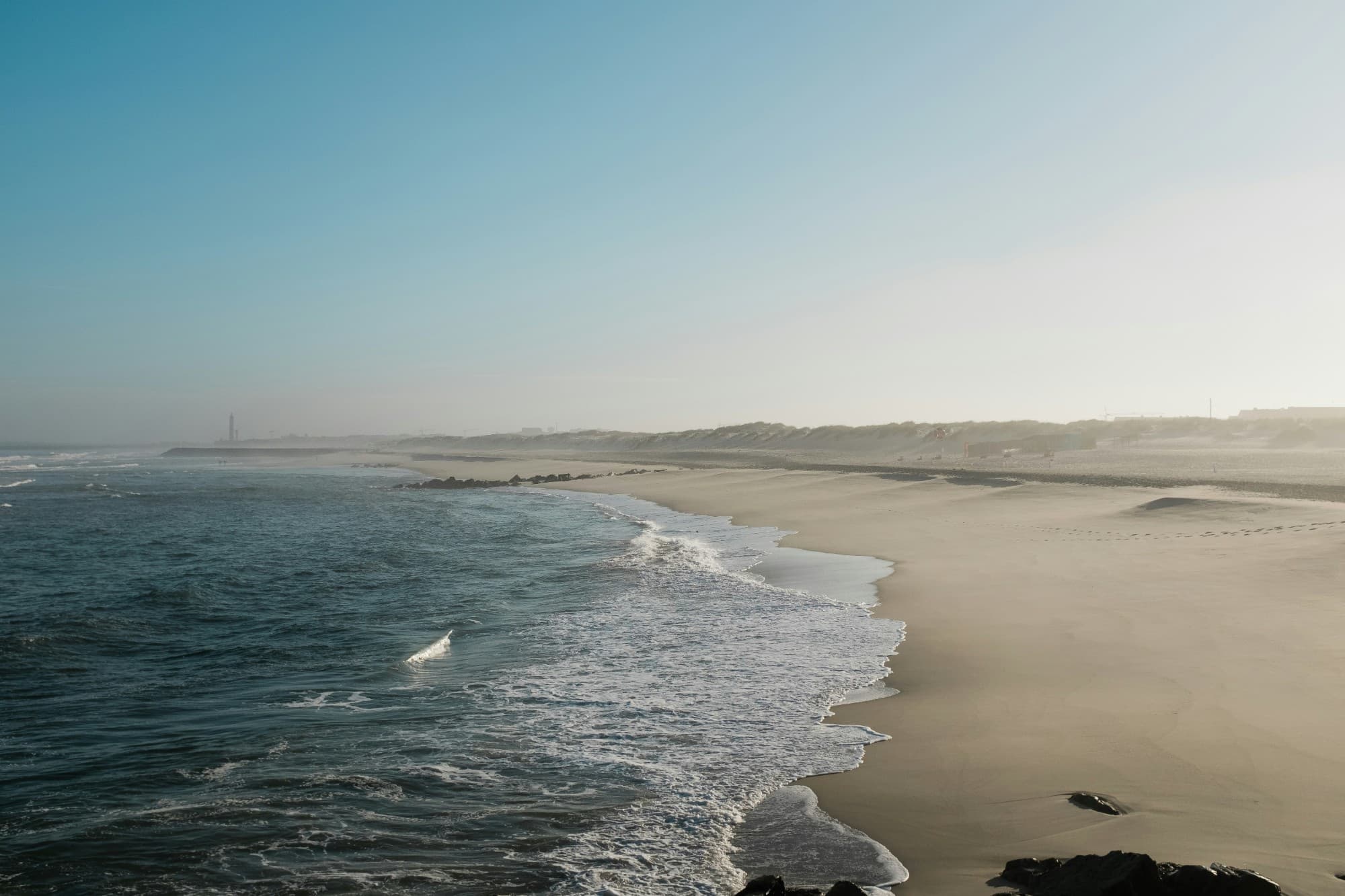 A wide, empty Atlantic beach with breaking waves and the Barra lighthouse in the haze